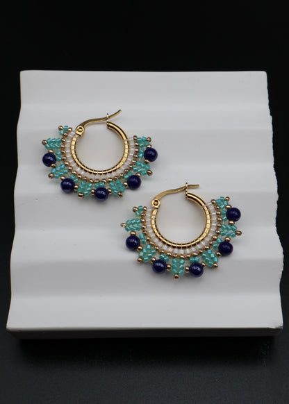 Gold hoop earrings with blue and green beads on a white stand.