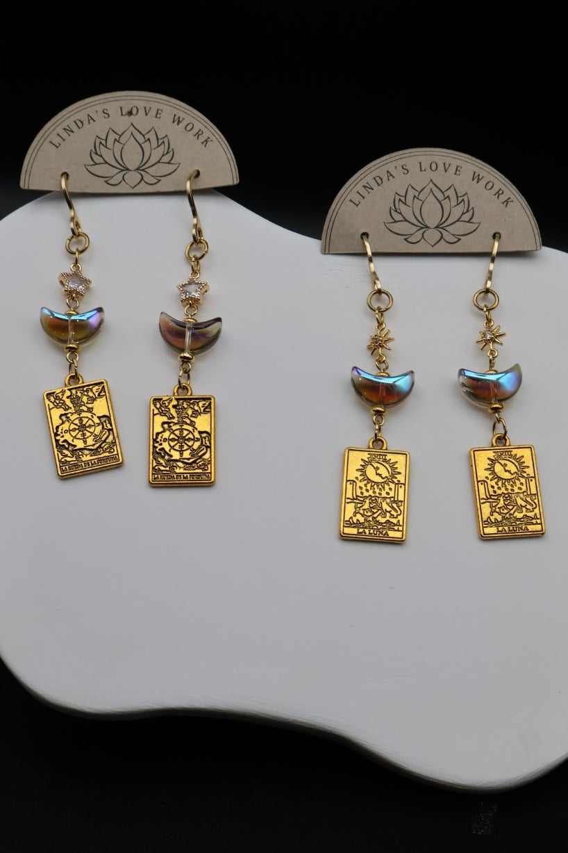 A pair of gold-toned dangle earrings displayed on a white stand against a black background. Each earring has a small faceted bead, a crescent moon charm, and a rectangular engraved plate with the words 'LOVE WORK' and a lotus symbol.