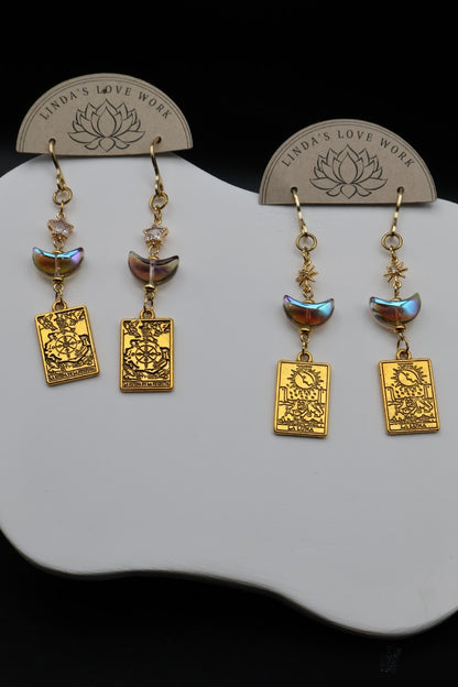 A pair of gold-toned dangle earrings displayed on a white stand against a black background. Each earring has a small faceted bead, a crescent moon charm, and a rectangular engraved plate with the words 'LOVE WORK' and a lotus symbol.