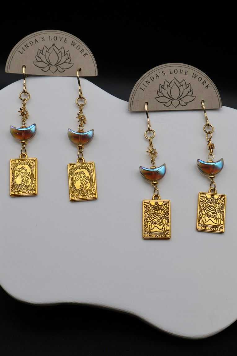 A pair of gold-toned dangle earrings displayed on a white stand against a black background. Each earring has a small iridescent blue-green bead, a crescent moon charm, and a rectangular gold pendant with an embossed profile.