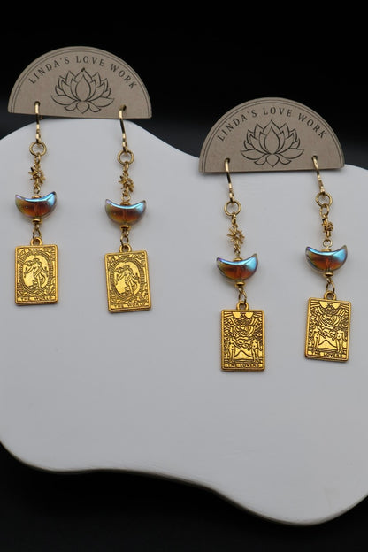 A pair of gold-toned dangle earrings displayed on a white stand against a black background. Each earring has a small iridescent blue-green bead, a crescent moon charm, and a rectangular gold pendant with an embossed profile.