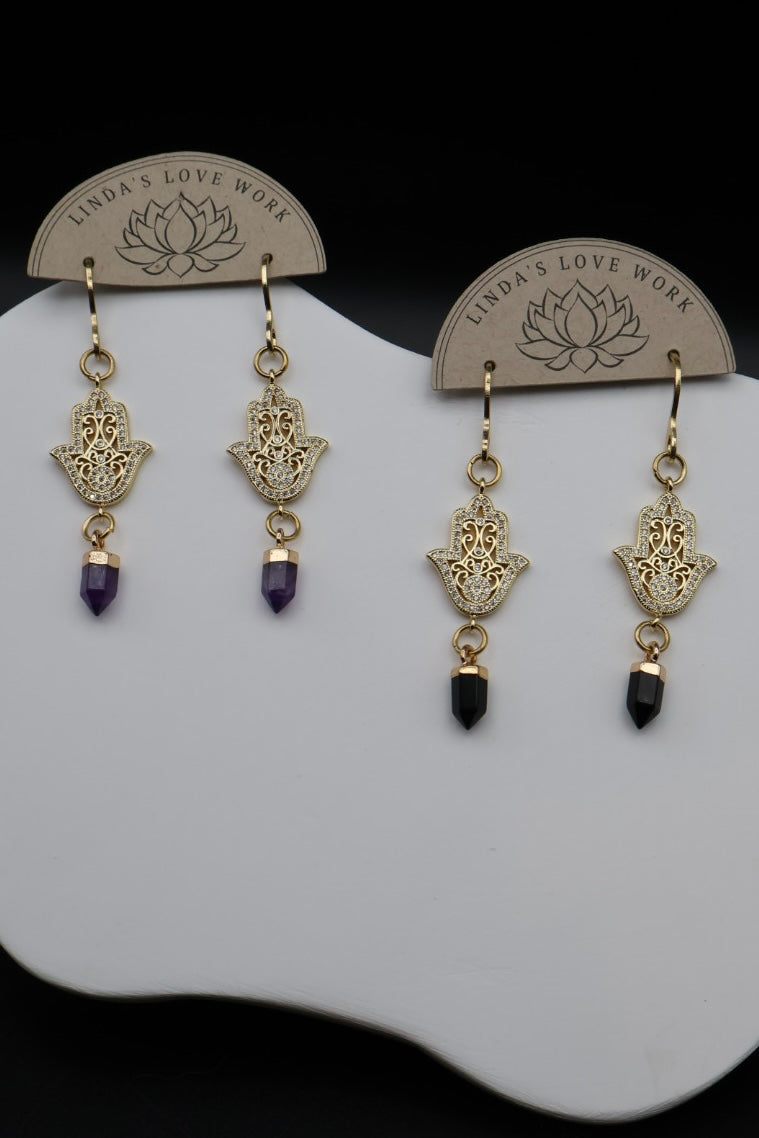 A pair of gold-tone Hamsa hand dangle earrings with purple amethyst and black obsidian beads, displayed on a white stand with branded backing cards.