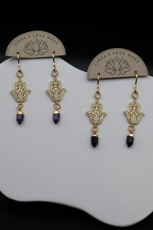 A pair of gold-tone Hamsa hand dangle earrings with purple amethyst and black obsidian beads, displayed on a white stand with branded backing cards.