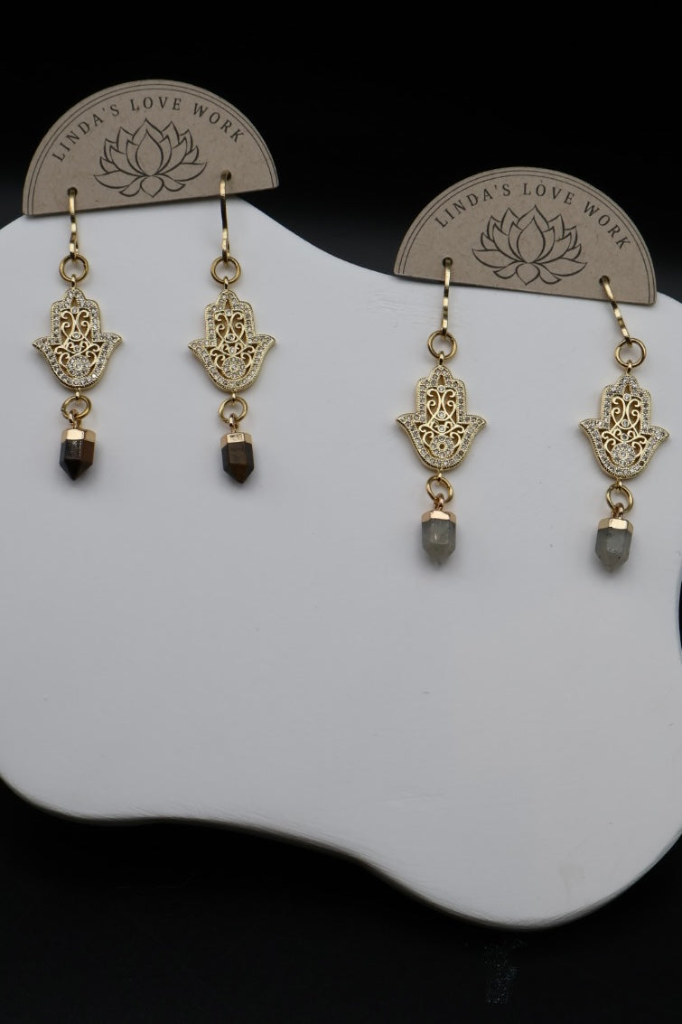 A pair of gold Hamsa hand dangle earrings with dark brown and gray bead drops, displayed on a white stand with brand card inserts.