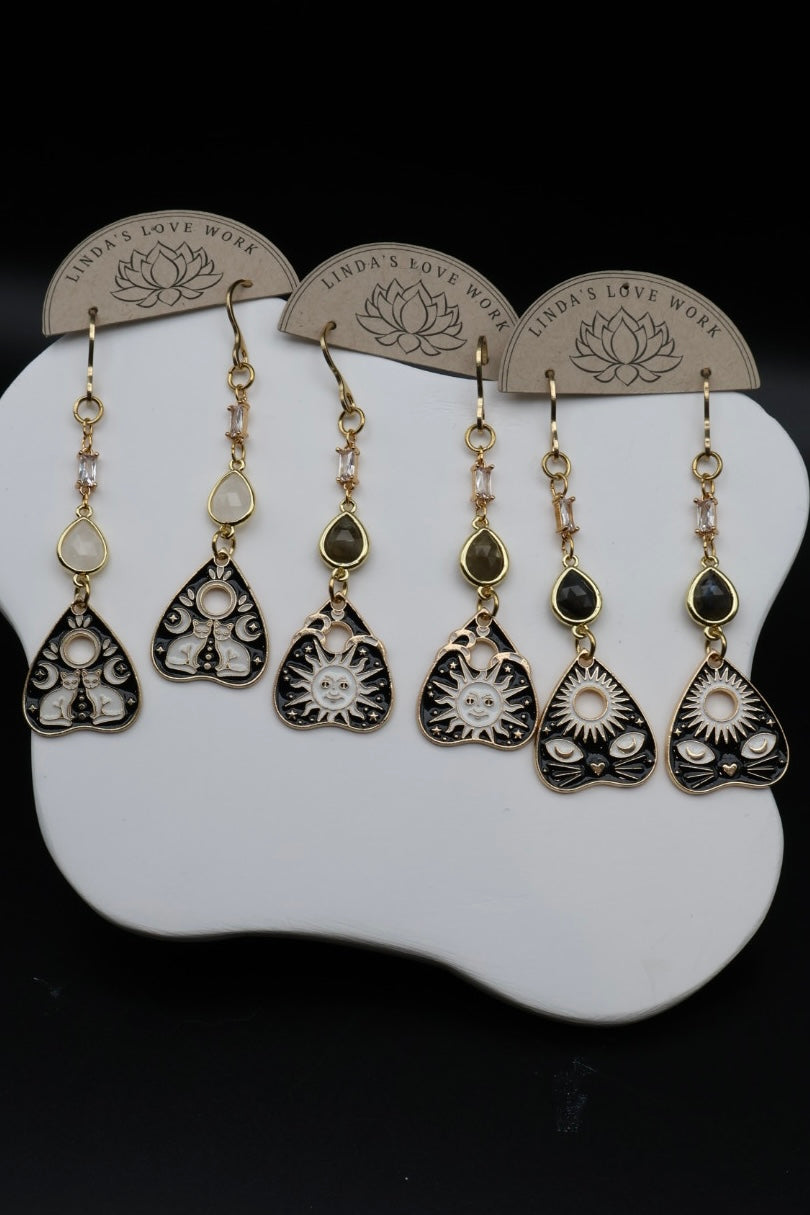 Five pairs of bohemian dangle earrings displayed on a white sculptural stand against a black background. The earrings feature silver and gold-toned metal, clear crystals, and dark green gemstone accents, with three pairs hanging from branded paper clips engraved with 'LINDA'S LOVE WORK'.