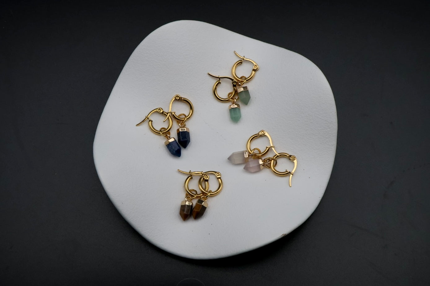 Four gold hoop earrings with dangling gemstone charms in blue, green, pink, and brown, arranged on a white ceramic dish against a dark background.