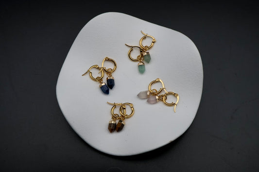 Four gold hoop earrings with dangling gemstone charms in blue, green, pink, and brown, arranged on a white ceramic dish against a dark background.
