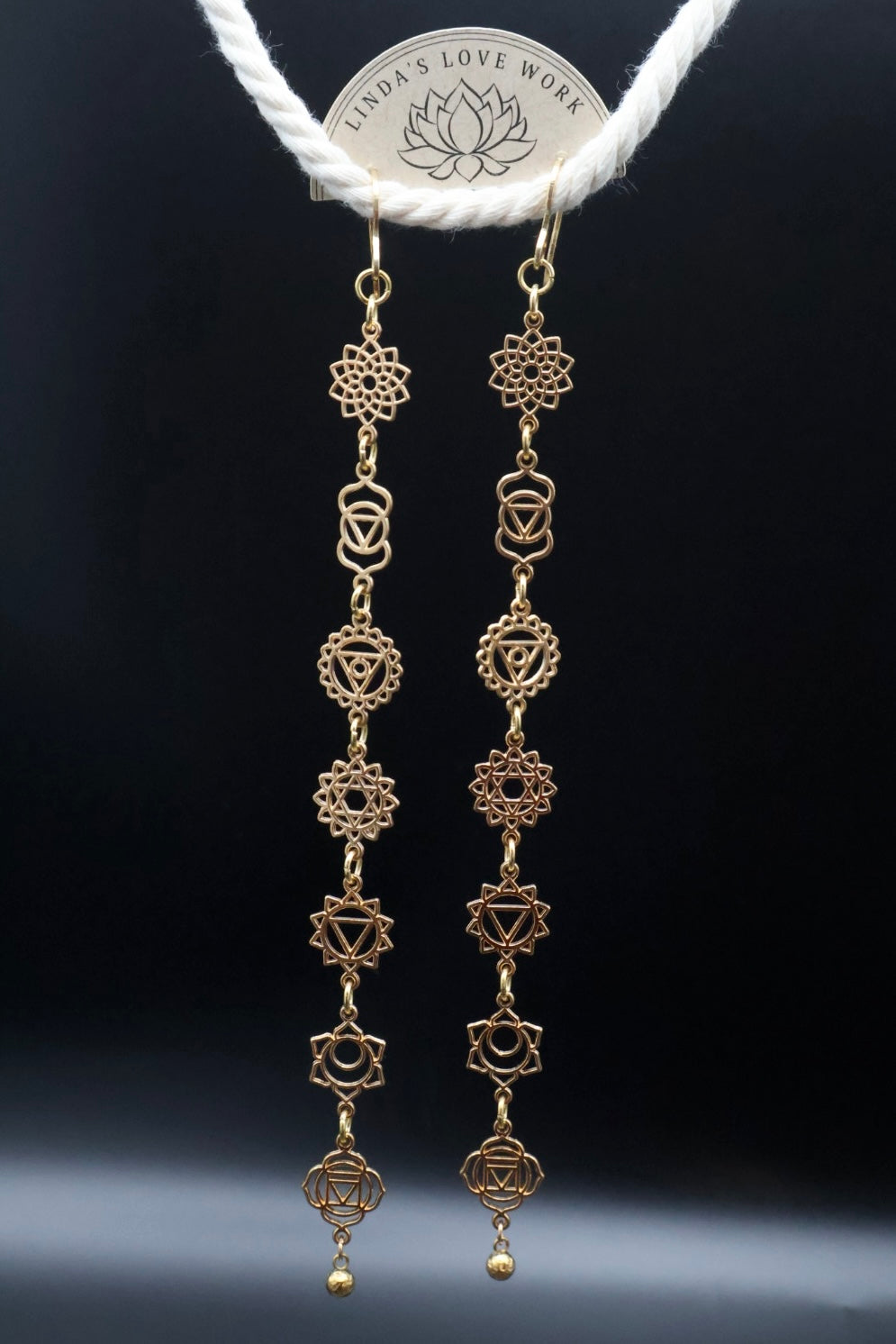 A pair of gold-toned dangle earrings with multiple chakra and geometric symbols, hanging from a white rope with a coin at the top against a dark background.