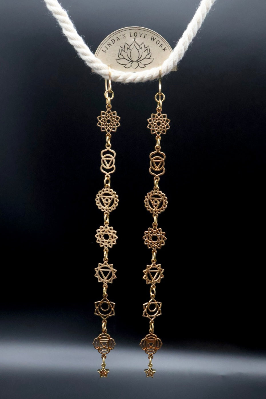 A pair of gold-toned dangle earrings with multiple chakra symbols, hanging from a white rope display. The display tag at the top reads 'Tender's Love Work'.