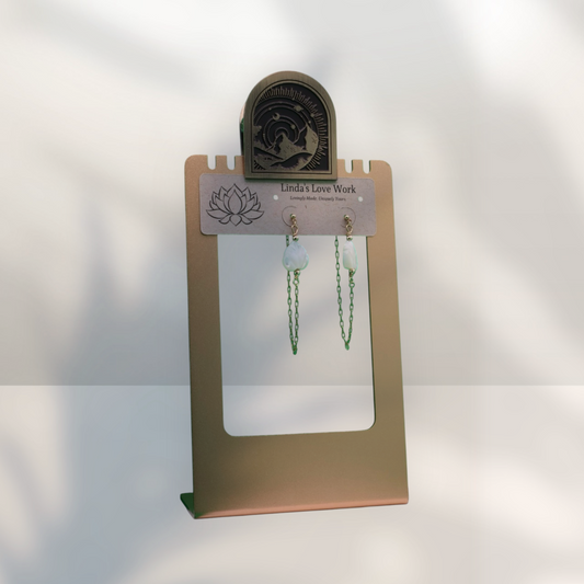 A pair of green beaded chain earrings displayed on a gold-colored jewelry stand with a sign that reads 'Linda's Love Work'.