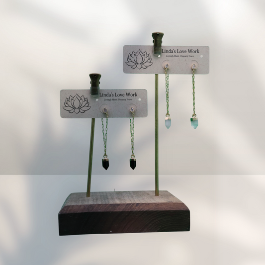 Two pairs of dangling earrings displayed on a wooden stand with branded cards reading 'Linda's Love Work.' The earrings have green chains, small silver-toned studs, and one black bead drop and one light blue bead drop.
