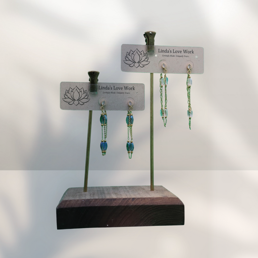 Two pairs of dangling beaded earrings displayed on small stand cards mounted on a wooden base. The earrings feature blue and green beads with silver-toned metal findings.