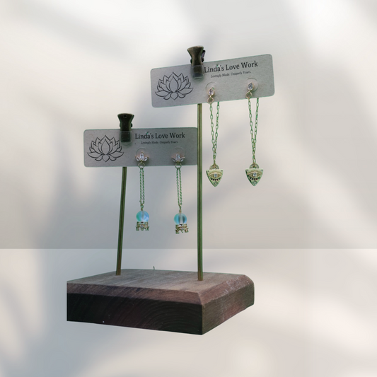 Two metal display stands holding three pairs of dangling earrings, photographed against a soft light gray background with leaf shadows. The stands are branded Linda's Love Work.