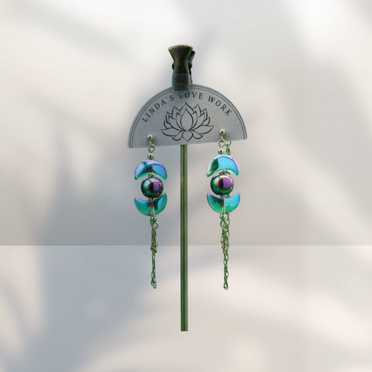A pair of dangling earrings displayed on a metal stand against a soft light gray background. The earrings feature iridescent teal-green disc-like beads with a central purple and green marbled bead, hanging from gold-toned chains.