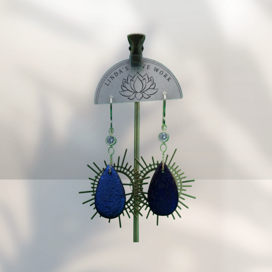 A pair of handmade dangle earrings displayed on a green stand. The earrings feature textured, teardrop-shaped dark blue pearly beads surrounded by green spiky wire details, with small clear faceted beads above.