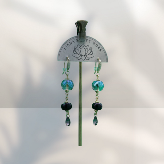 A pair of dangling earrings displayed on a metal stand. The earrings feature faceted teal-blue beads, small silver-toned chain links, a dark black bead, and a teardrop-shaped blue-green drop.