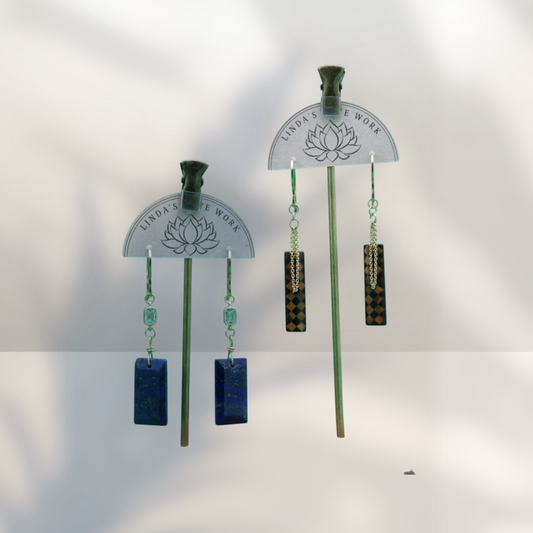 Two pairs of Linda's Love Work dangle earrings displayed on metal stands against a white background with soft shadows. One pair features rectangular blue stones, and the other features patterned brown and black rectangular stones with light blue beads.