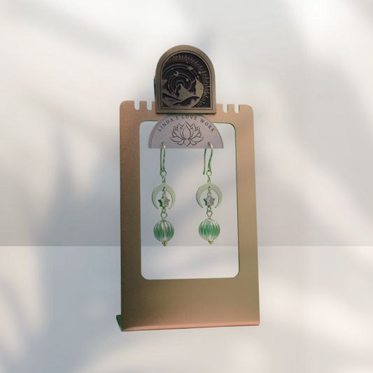 A pair of green beaded dangle earrings displayed on a metallic gold stand with a small arched decorative plaque at the top. The earrings have circular green elements and bead drops, shown against a softly lit white background with leaf shadows.