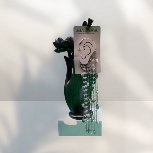 A pair of long dangling earrings displayed on a card labeled Linda's Little Work, hanging in front of a dark green vase against a white background with soft shadows.