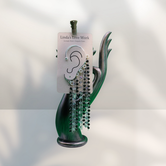 A pair of dangling beaded earrings displayed on a white card labeled 'Linda's Love Work,' mounted on a green sculptural stand against a softly lit white background.