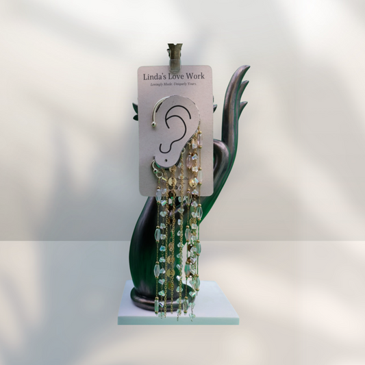 A pair of long dangling earrings displayed on a white backing card labeled 'Linda's Love Work,' mounted on a sculptural black stand against a softly lit white background.