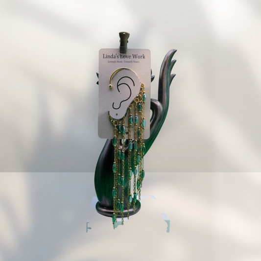 A pair of gold hoop earrings with long dangling chains and green faceted beads, displayed on a card labeled Linda's Lave Work, set against a dark green sculptural stand.