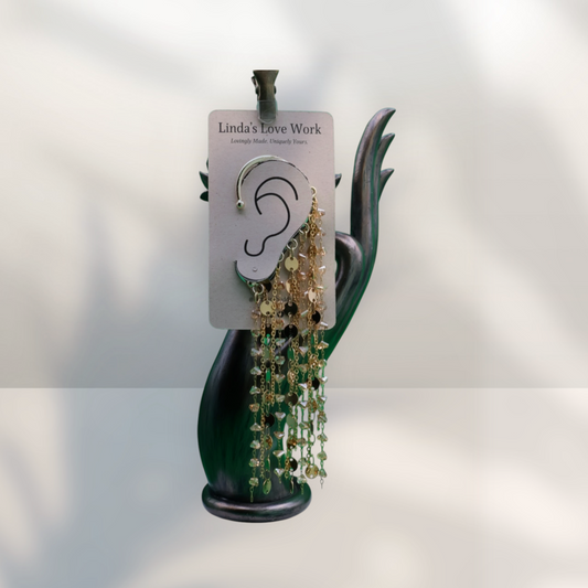 A pair of dangling hoop earrings with green and gold beaded chains, displayed on a white card labeled 'Linda's Love Work' and mounted on a dark green hand-shaped stand against a light gray background.