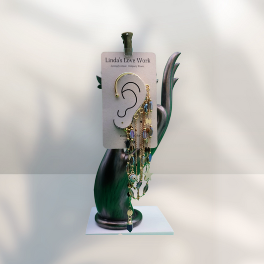 A pair of dangling gold-toned earrings with blue and green faceted beads displayed on a branded card stand shaped like a hand, set against a softly lit white background.