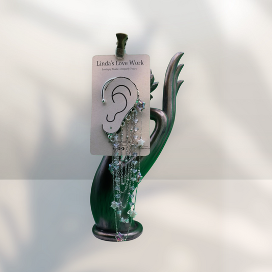 A pair of silver hoop earrings with dangling beaded chains and floral charms, displayed on a white card labeled 'Linda's Love Work' and held in a dark green hand-shaped jewelry stand against a softly lit white background.