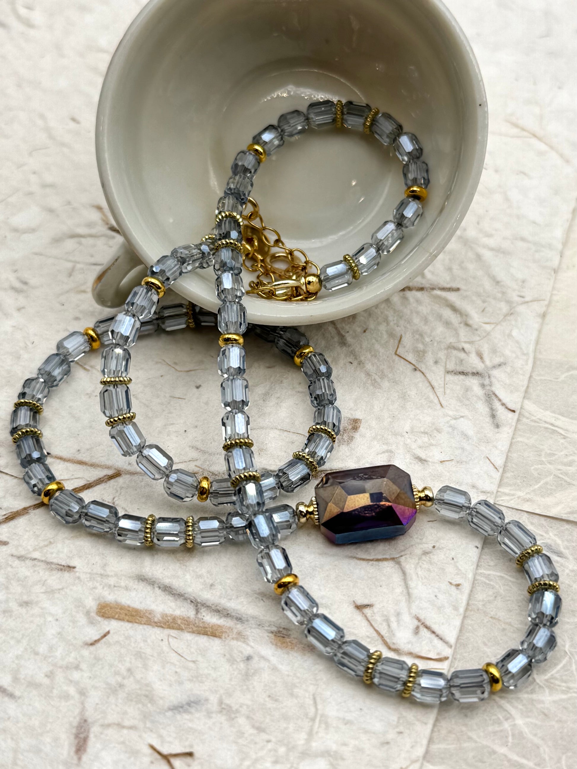 Beaded necklace with a large purple bead on a textured surface with a cup in the background.