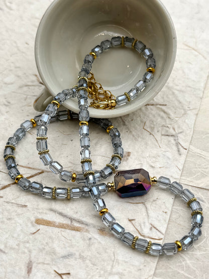 Beaded necklace with a large purple bead on a textured surface with a cup in the background.
