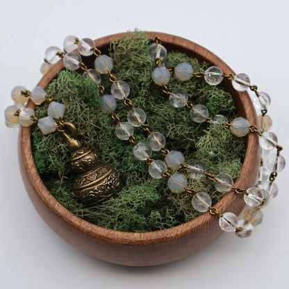 Decorative necklace with clear beads and a bronze pendant on a bed of green moss in a wooden bowl.