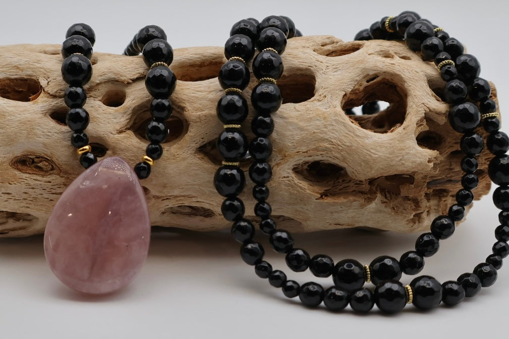 Black beaded necklace with a pink pendant on a piece of driftwood.