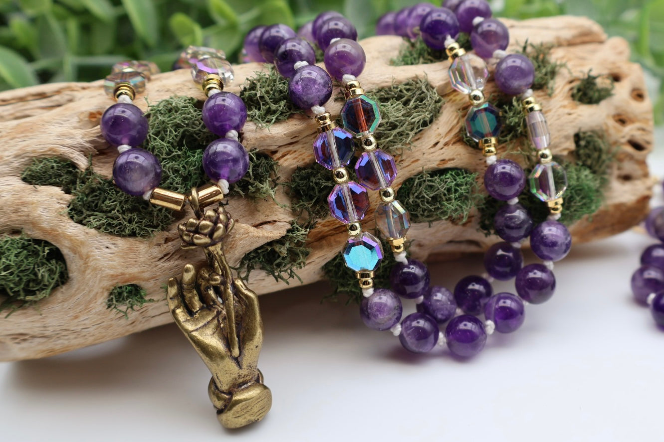 Purple beaded bracelets with a bronze hand charm on a piece of driftwood.