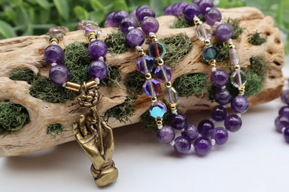 Purple beaded bracelets with a bronze hand charm on a piece of driftwood.