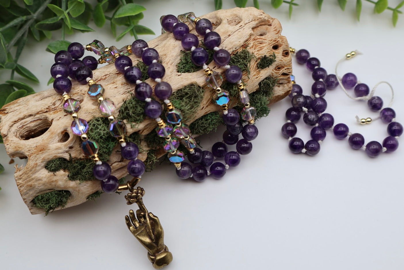 Purple beaded jewelry set with a gold hand charm on a white surface with greenery.