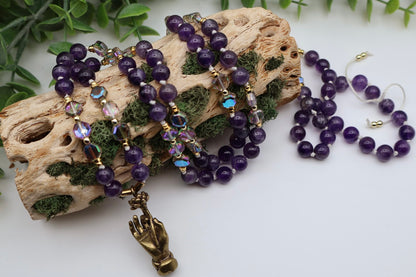 Purple beaded jewelry set with a gold hand charm on a white surface with greenery.