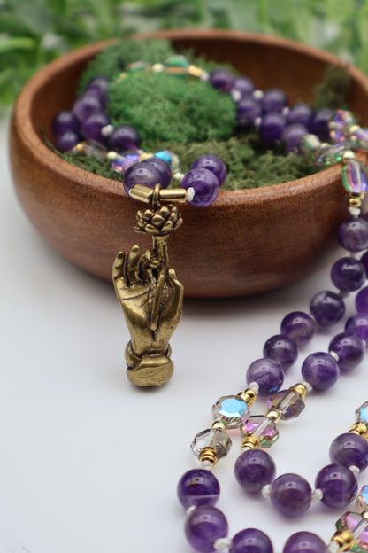 Purple beaded necklace with a gold hand pendant on a white surface with a wooden bowl and greenery in the background.