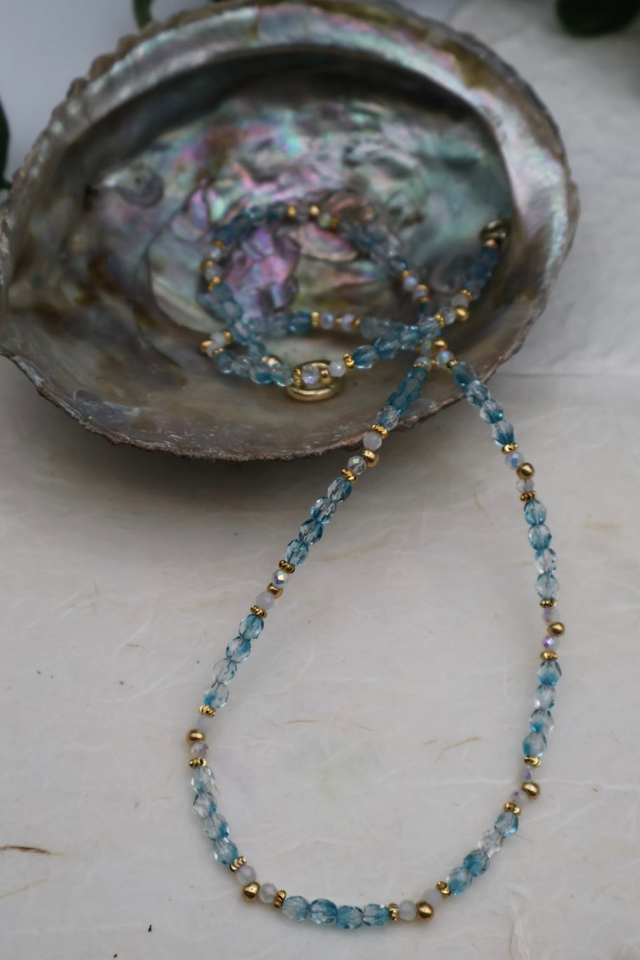 Blue beaded necklace with gold accents on a white surface with an open shell in the background.