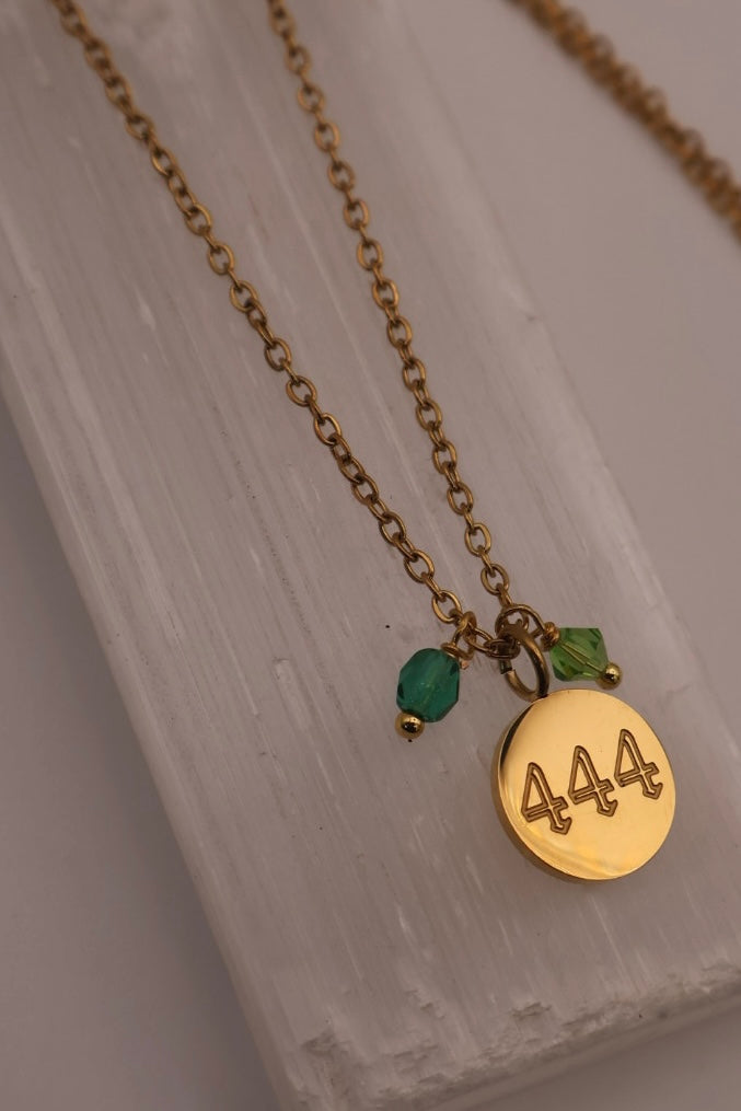 Gold necklace with a '444' pendant and green beads on a light wooden surface.