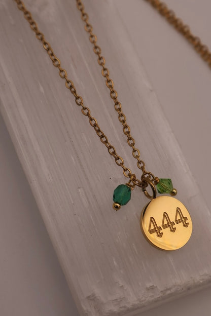 Gold necklace with a '444' pendant and green beads on a light wooden surface.