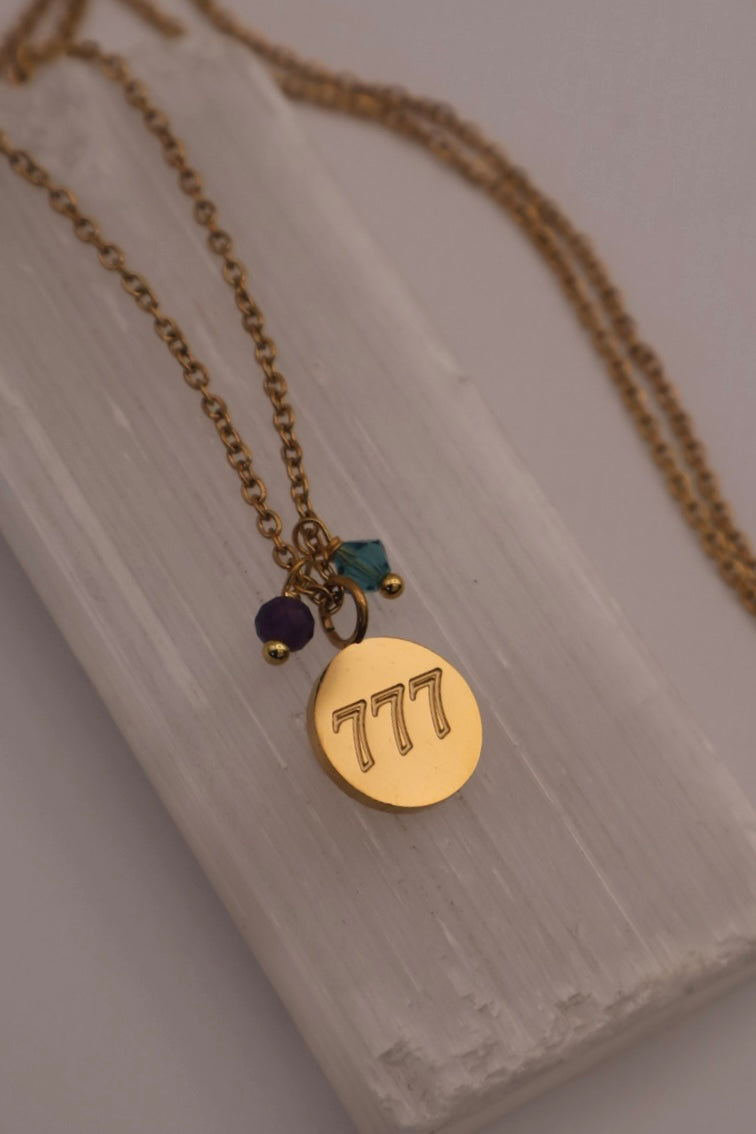 Gold necklace with a round pendant featuring the number '777' on a light wooden surface.