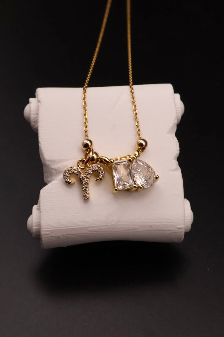 A gold chain necklace with a triple charm pendant, featuring a gold zodiac symbol, a small clear gemstone, and a larger irregular clear gemstone, displayed on a white cylindrical stand against a black background.