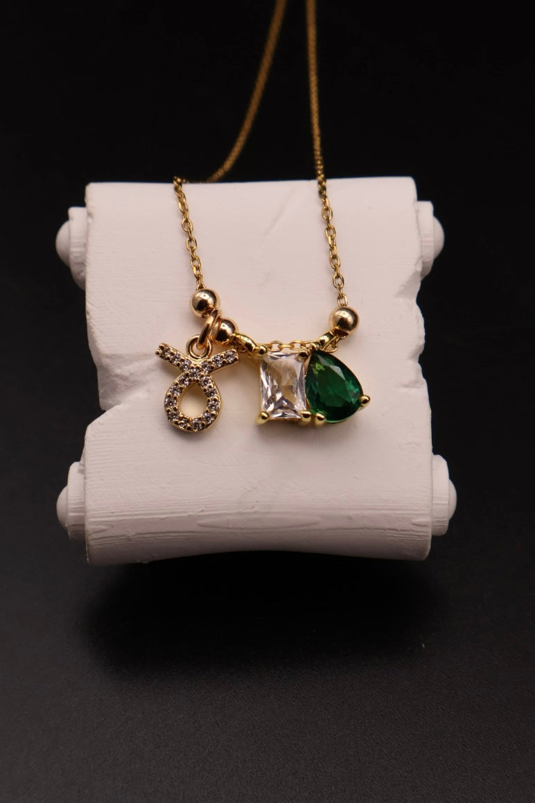 A gold chain necklace with three charms displayed on a white cylindrical stand against a black background. The charms include a textured gold zodiac sign, a clear square gemstone, and a green teardrop gemstone.