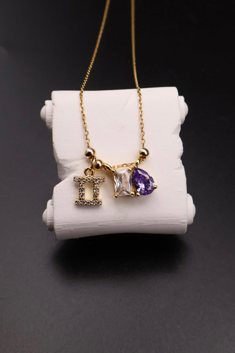 Gold necklace with Gemini zodiac symbol and amethyst teardrop charms, displayed on a white cylindrical stand against a dark background.