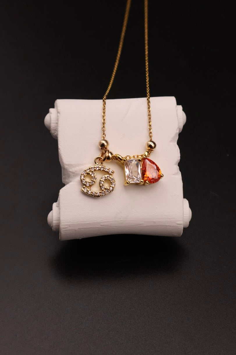 A gold necklace with three charms displayed on a white cylindrical stand against a black background. The charms include a gold infinity symbol, a clear faceted gemstone, and a small orange-red teardrop gemstone.