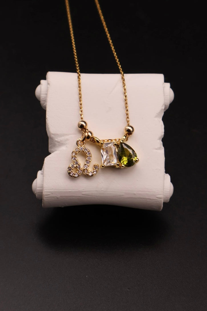 A gold chain necklace with three charms displayed on a white cylindrical stand against a black background. The charms include a gold zodiac sign, a clear faceted gemstone, and a green heart-shaped gemstone.