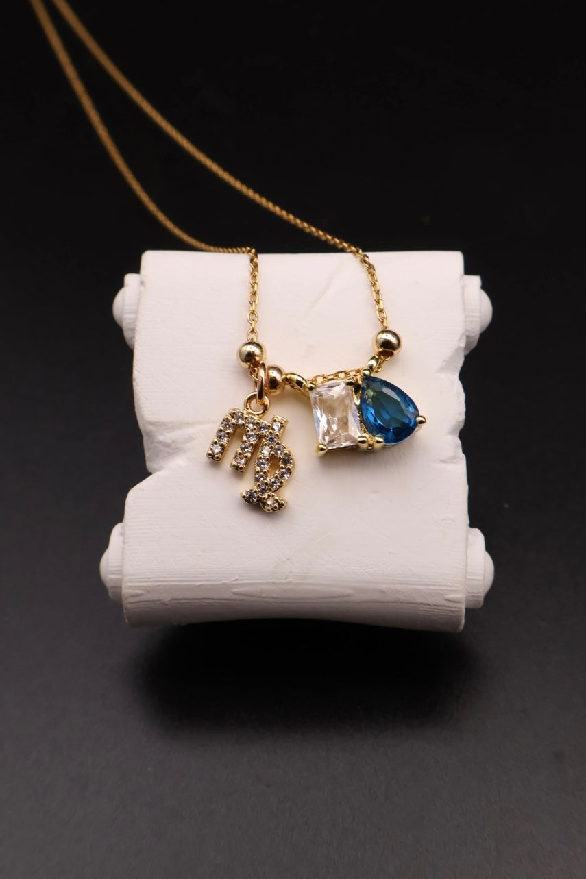 A gold chain necklace with a double pendant display on a white cylindrical stand. One pendant is a gold monogram letter 'M' set with small clear stones, and the other is a gold pendant featuring a clear rectangular stone and a blue teardrop-shaped gemstone, set against a dark background.