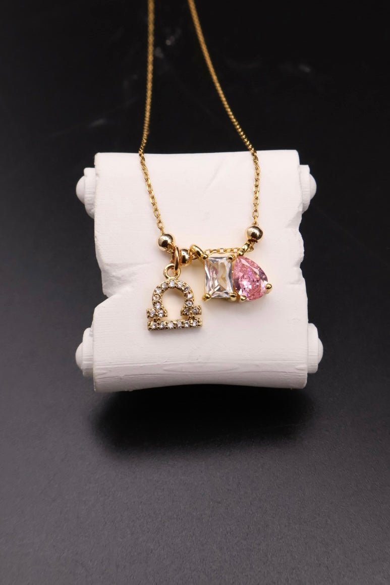 A gold chain necklace with three charms displayed on a white cylindrical stand against a dark background. The charms include a small gold heart with tiny clear stones, a square clear stone, and a teardrop-shaped pink gemstone.