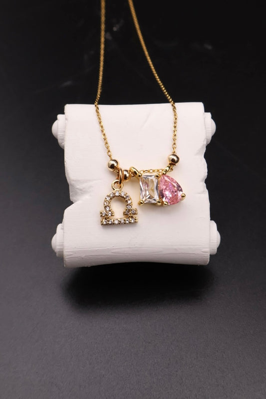 A gold chain necklace with three charms displayed on a white cylindrical stand against a dark background. The charms include a small gold heart with tiny clear stones, a square clear stone, and a teardrop-shaped pink gemstone.